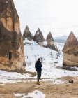 Назад погляд людини в темному теплому одязі, що дивиться в далечінь, стоячи на брудній дорозі проти снігового пагорба з відомими стовпами з гострими вершинами у вигляді списа в національному парку в Туреччині. — стокове фото