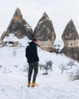 Vue arrière de l'homme en vêtements chauds sombres regardant loin tout en se tenant sur la route de terre contre la colline enneigée avec des piliers célèbres avec des sommets pointus en forme de lance dans le parc national en Turquie — Photo de stock