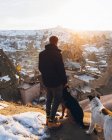 Vista trasera de un joven hombre irreconocible con ropa de abrigo de pie acariciando perros en la colina contra pequeñas antiguas casas cueva en el valle nevado al atardecer en Turquía - foto de stock