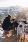 Молодой неузнаваемый человек в теплой одежде ласкает собак на холме против маленьких древних пещерных домиков в заснеженной долине в сумерках в Турции — стоковое фото