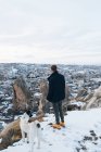 Обратный вид молодого неузнаваемого человека в теплой одежде, стоящего с верной собакой на холме напротив маленьких древних пещерных домиков в заснеженной долине в сумерках в Турции — стоковое фото