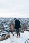 Vue arrière d'un jeune homme en vêtements chauds debout avec un chien loyal sur la colline contre de petites maisons de grotte anciennes dans la vallée enneigée au crépuscule en Turquie — Photo de stock