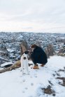 Visão traseira de um jovem agachado em roupas quentes com cão leal na colina contra pequenas casas de cavernas antigas no vale nevado ao anoitecer na Turquia — Fotografia de Stock