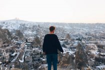 Rückansicht eines nicht wiederzuerkennenden jungen Mannes in warmer Kleidung, der auf einem schneebedeckten Hügel steht und über eine berühmte Kleinstadt mit alten Höhlenhäusern im Tal bei Sonnenuntergang in der Türkei blickt — Stockfoto