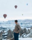 Seitenansicht eines nicht wiederzuerkennenden Mannes, der bei trübem Wetter in der Türkei vor ungewöhnlichen Steinsäulen und bunten Luftballons steht, die in den Himmel über nebligem, verschneitem Hochland rasen — Stockfoto