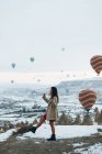 Вид сбоку женщины в теплой одежде, смотрящей вверх, стоя против необычных каменных столбов и красочных воздушных шаров, мчащихся в небе над туманным снежным нагорьем в пасмурную погоду в Турции — стоковое фото