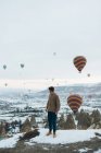 Боковой вид неузнаваемого человека, смотрящего на пейзаж, стоя против необычных каменных столбов и красочных воздушных шаров, мчащихся в небе над туманной снежной горной местностью в пасмурную погоду в Турции — стоковое фото