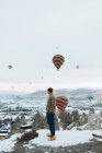 Side view of unrecognizable man looking away at landscape while standing against unusual stone pillars and colorful air balloons racing in sky over foggy snowy highland in overcast weather in Turkey — Stock Photo