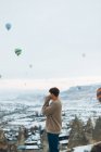 Side view of unrecognizable man looking away at landscape while standing against unusual stone pillars and colorful air balloons racing in sky over foggy snowy highland in overcast weather in Turkey — Stock Photo