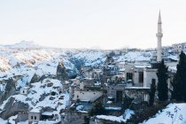Desde arriba pequeña ciudad con edificios antiguos en valle y alta torre de minarete en la ladera nevada contra el cielo azul sin nubes en invierno en Turquía - foto de stock