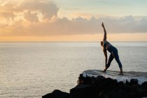 Невпізнавана жінка на спортивному лаві стоїть у позі Триконасани на йога маті проти розореного океану під хмарним небом на заході сонця. — стокове фото