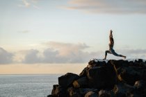 Vista lateral de una mujer anónima practicando yoga con los brazos levantados sobre rocas contra el océano bajo el cielo nublado al atardecer - foto de stock