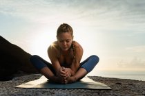 Erdgeschoss einer jungen Frau mit geschlossenen Augen, die Beine und Rücken streckt, während sie Yoga an der Meeresküste bei Sonnenschein praktiziert — Stockfoto