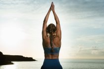 Rückansicht einer nicht wiederzuerkennenden Frau, die in Vrksasana-Pose steht, während sie abends Yoga auf Felsbrocken gegen den wogenden Ozean praktiziert — Stockfoto
