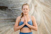 Joven mujer consciente en ropa deportiva meditando con los ojos cerrados mientras practica yoga sobre fondo borroso - foto de stock