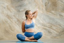 Joven hembra sentada mirando hacia otro lado estirando los brazos mientras practica yoga en la esterilla contra el monte - foto de stock
