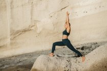 Junge barfüßige Frau in Sportbekleidung praktiziert Yoga auf Halbmond Pose gegen felsigen Berg im Sonnenlicht — Stockfoto