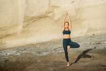 Молодая внимательная женщина в спортивной одежде на vrksasana с закрытыми глазами, практикуя йогу стоя на скалистом фоне — стоковое фото