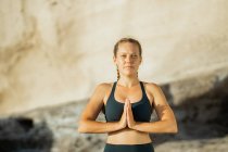 Joven mujer consciente en ropa deportiva meditando mirando a la cámara mientras practica yoga sobre fondo borroso - foto de stock
