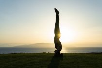 Side view of anonymous female silhouette standing on head while practicing yoga on ocean coast in sunlight at dusk — Stock Photo