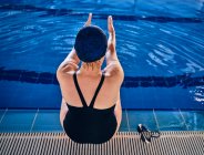 Back view high angle of anonymous female swimmer doing side bend exercise while sitting at poolside and stretching arms before water aerobics — Stock Photo