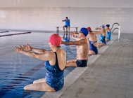 Group of people in swimwear sitting at poolside and stretching raised arms while exercising during water aerobics training with instructor in pool — Stock Photo