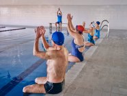 Group of people in swimwear sitting at poolside and stretching raised arms while exercising during water aerobics training with instructor in pool — Stock Photo
