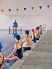Gruppo di persone in costume da bagno sedute a bordo piscina e che allungano le braccia sollevate durante l'allenamento di acquagym con istruttore in piscina — Foto stock