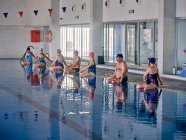 Grupo de personas en traje de baño sentadas junto a la piscina y estirando los brazos levantados mientras hacen ejercicio durante el entrenamiento de aeróbic acuático - foto de stock