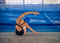 Вид ззаду високий кут анонімного плавця, який виконує бічні вправи згинання, сидячи біля басейну та розтягуючи руки перед водною аеробікою — стокове фото