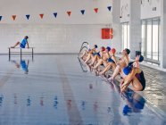 Grupo de pessoas em trajes de banho sentado à beira da piscina e alongamento braços levantados durante o exercício durante o treinamento de aeróbica aquática com instrutor na piscina — Fotografia de Stock