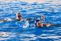 Sonrientes hembras de mediana edad en traje de baño nadando en la piscina y haciendo ejercicio durante aeróbic acuático juntas - foto de stock