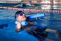 Vista lateral do macho de meia-idade em óculos nadando de volta no macarrão de espuma durante a aeróbica aquática na piscina — Fotografia de Stock
