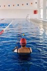 Desde arriba vista trasera de una anciana nadadora anónima con gorra de pie en el agua de la piscina - foto de stock