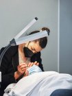 Esteticista profissional tratando pestanas de paciente feminino em máscara protetora no salão de beleza moderno com lâmpada de suporte — Fotografia de Stock