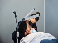 Professionelle Kosmetikerin behandelt Wimpern einer Patientin in Schutzmaske im modernen Schönheitssalon mit Standlampe — Stockfoto