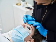 Cosmetician profissional irreconhecível cortado em luvas de látex usando pinças ao modelar sobrancelhas de paciente fêmea durante o procedimento de beleza no salão — Fotografia de Stock