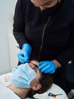 Fechar a cultura cosmetologista anônimo em luvas de látex usando pincel curler cílios durante o procedimento de beleza para o cliente feminino — Fotografia de Stock