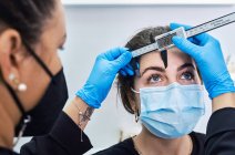 Esthéticienne cultivée méconnaissable dans des gants en latex à l'aide d'une règle pour mesurer les sourcils du client féminin dans un masque de protection obtenir une procédure de beauté pendant la pandémie de coronavirus — Photo de stock