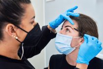 Beautician in latex gloves brushing eyebrow of young female client in protective mask during beauty appointment in modern salon — Stock Photo