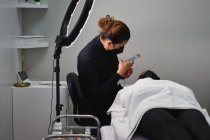 Cosmetologist profissional com smartphone tirando foto do rosto do cliente feminino recebendo tratamento de cílios durante o procedimento de beleza no salão — Fotografia de Stock