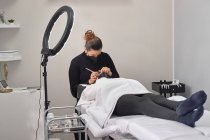 Professionelle Kosmetikerin mit Pinzette behandelt Wimpern weiblicher Kunden mit Gesichtsmaske während der Wimpernverlängerung im modernen Schönheitssalon mit Ringlicht-Lampe — Stockfoto
