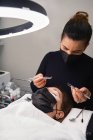 Cosmetician profissional com pinças que tratam pestanas do cliente fêmea com máscara facial durante o procedimento de extensão de pestanas no salão de beleza moderno com lâmpada de luz de anel — Fotografia de Stock