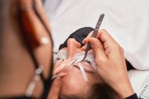Professionelle Kosmetikerin mit Pinzette behandelt Wimpern weiblicher Kunden mit Gesichtsmaske während der Wimpernverlängerung im modernen Schönheitssalon mit Ringlicht-Lampe — Stockfoto