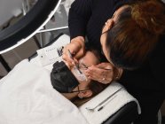 Сверху профессиональный косметик с пинцетом лечит ресницы клиентки с маской для лица во время процедуры наращивания ресниц в современном салоне красоты с кольцевой лампой — стоковое фото