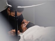 Cosmético profesional con pinzas que tratan las pestañas de cliente femenino con máscara facial durante el procedimiento de extensión de pestañas en el salón de belleza moderno con lámpara de luz de anillo - foto de stock