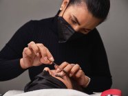 Primo piano di coltura cosmetologo anonimo in guanti di lattice utilizzando pennello bigodino ciglia durante la procedura di bellezza al cliente femminile — Foto stock