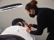 Cosmetologist profissional com o processo de tiroteio de telefone celular do procedimento de extensão de pestanas no salão de beleza com luz de anel — Fotografia de Stock