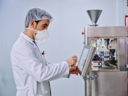Vista lateral del químico masculino en la máquina de llenado de cápsulas de operación uniforme en la planta de fabricación farmacéutica - foto de stock