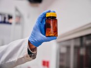 Crop scientifique anonyme dans des gants de protection debout montrant pile de capsules sur la bouteille en laboratoire — Photo de stock
